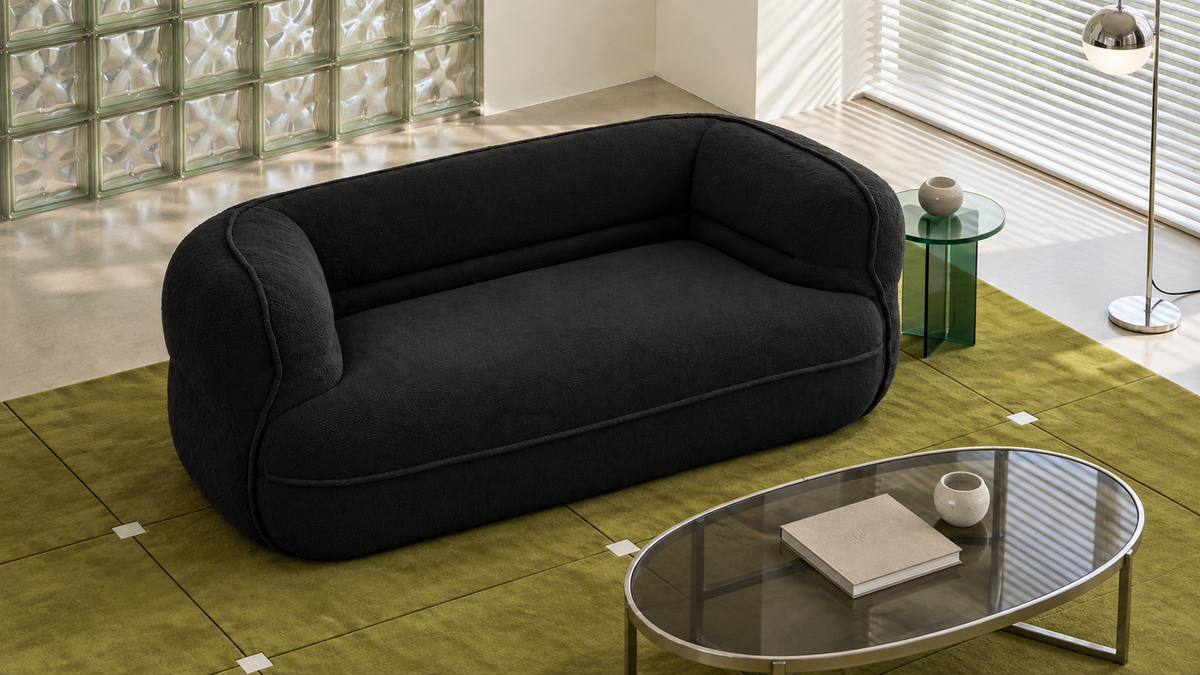 A modern black upholstered loveseat with rounded arms and a low back, shown in a styled living room setting with a green rug, glass coffee table, and side table.